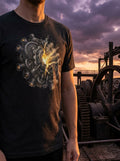 Person wearing a dark t-shirt with a mechanical design, standing in front of industrial machinery and a sunset sky.