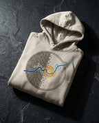 Oatmeal Heather Rewire hoodie folded on dark stone, showing the circular brown grid, blue current and gold spiral.