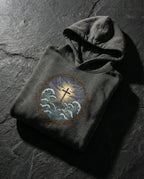 Charcoal Heather Trouble Hoodie folded on dark stone, showing cross above storm waves within a thorn border.