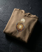 Latte Edify Hoodie folded on dark stone, showing interwoven golden figures rising toward a light.