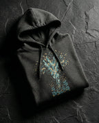 Charcoal Heather Thirty Sixty Hundred Hoodie folded on dark stone, showing a tall teal, navy and gold spray of seeds and leaves above small seedlings.