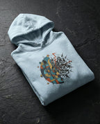Sky Blue Cease Striving Hoodie folded on dark stone, showing gold and blue interlocking forms beside scattered black fragments.