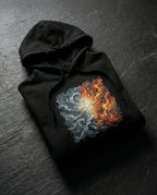 Black Unconsumed Hoodie folded on dark stone, showing blue waves and orange flames surrounding a glowing cross.