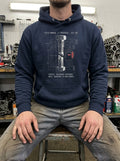 A man sitting in a gritty workshop wearing a navy blue Selah Supply Co. hoodie. The graphic features a technical blueprint of a fractured metal bolt and a red gauge. Text reads Component Torque Spec 119.105. Parable-based Christian streetwear.