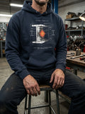 Man in a gritty industrial workshop wearing a Selah Supply Co Navy Blazer 'Component: Yield' hoodie featuring a technical I-beam heat treatment graphic inspired by 1 Peter 1:7.
