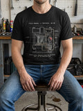 Man in a gritty industrial workshop wearing a Selah Supply Co Black Heather graphic tee featuring a hydraulic press schematic inspired by 2 Corinthians 4:8