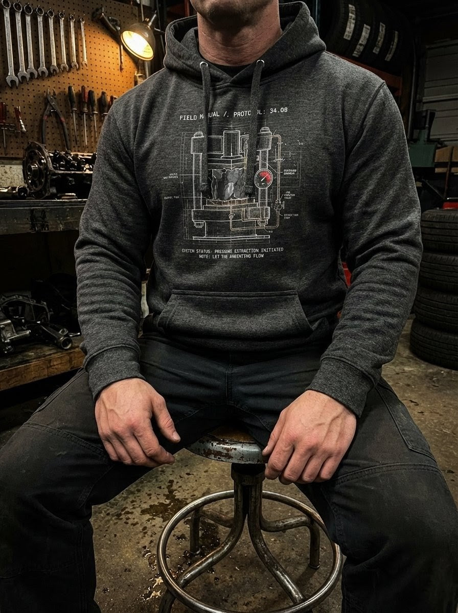 An operator in a rustic garage workshop wearing the charcoal Extraction hoodie. The chest graphic shows a detailed industrial schematic of a hydraulic press, representing the 'Story of the Load' from the Protocol Collection.