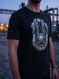 Front-facing lookbook photograph of a model wearing the Black Heather variant of the premium graphic t-shirt. The garment features a large, centered circular industrial emblem with intricate mechanical core lines, captured in an outdoor setting with warm, high-contrast sunset lighting reflecting off background industrial structures.