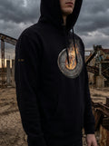 Model wearing the Selah Supply Co. Black Transmission: Firewall Falls // FREQ: 01.10 streetwear hoodie, showcasing the industrial brutalist circular graphic artwork against a rusty steel infrastructure background.