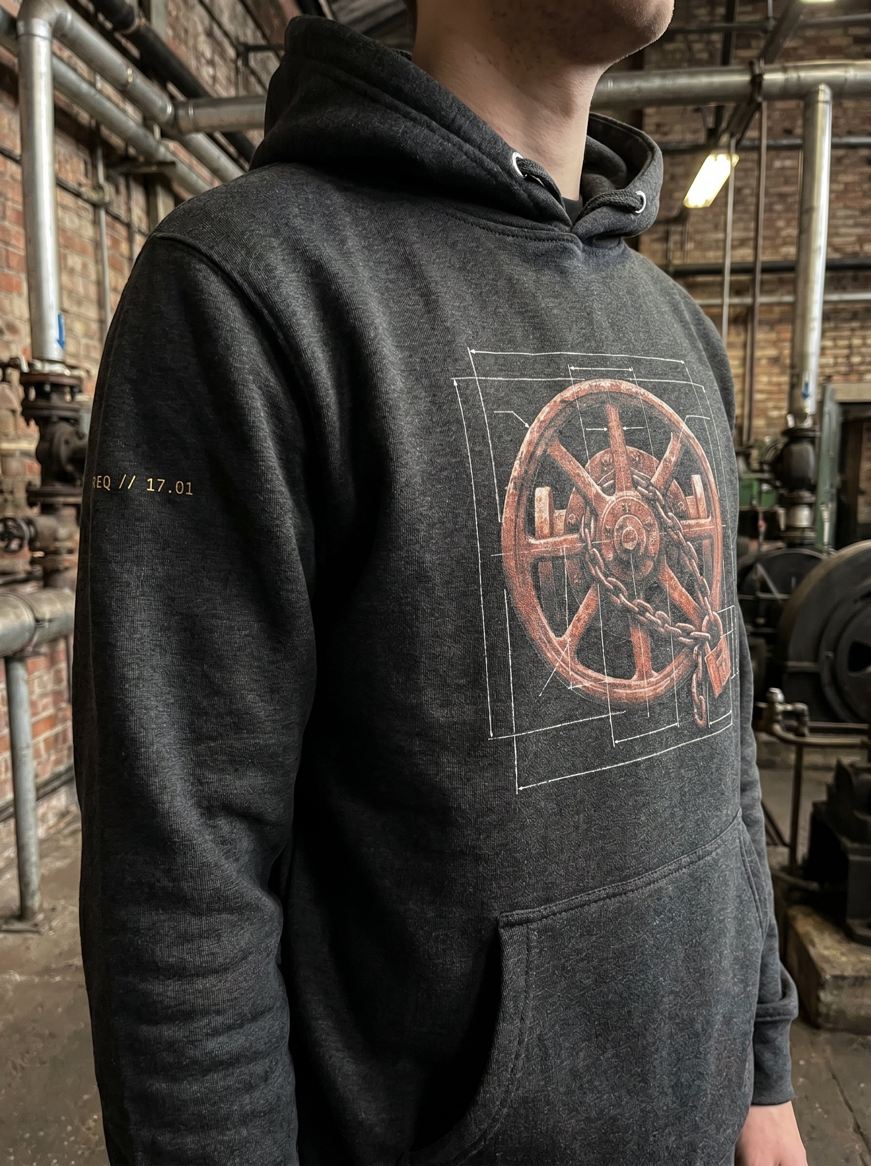Outdoor mid-range lookbook photograph of a model wearing the black industrial graphic hoodie. The shot captures a side-angle view of the garment, highlighting the scale of the graphic layout against a real-world background of structural steel scaffolding and weathered utility infrastructure under natural daylight.