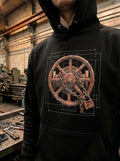 Front-facing vertical lookbook photograph of a model wearing a black premium graphic hoodie. The chest print features a prominent industrial graphic of a weathered wooden wheel bound by a heavy metal chain and padlock, layered over a white technical blueprint grid schematic. Set against an outdoor industrial backdrop of plumbing pipes.