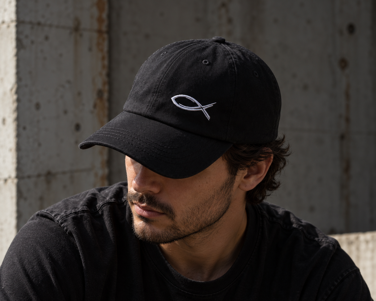A man wearing a black baseball cap with a white embroidered fish symbol, posed against a textured concrete wall.