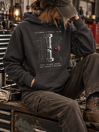 Model seated in a workshop wearing a Charcoal Heather Torque hoodie, face obscured by a cap, with the sheared-bolt graphic above the pouch pocket.