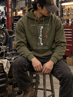 Model seated in a workshop wearing a Military Green Torque hoodie, face obscured by a cap, with the sheared-bolt graphic above the pouch pocket.