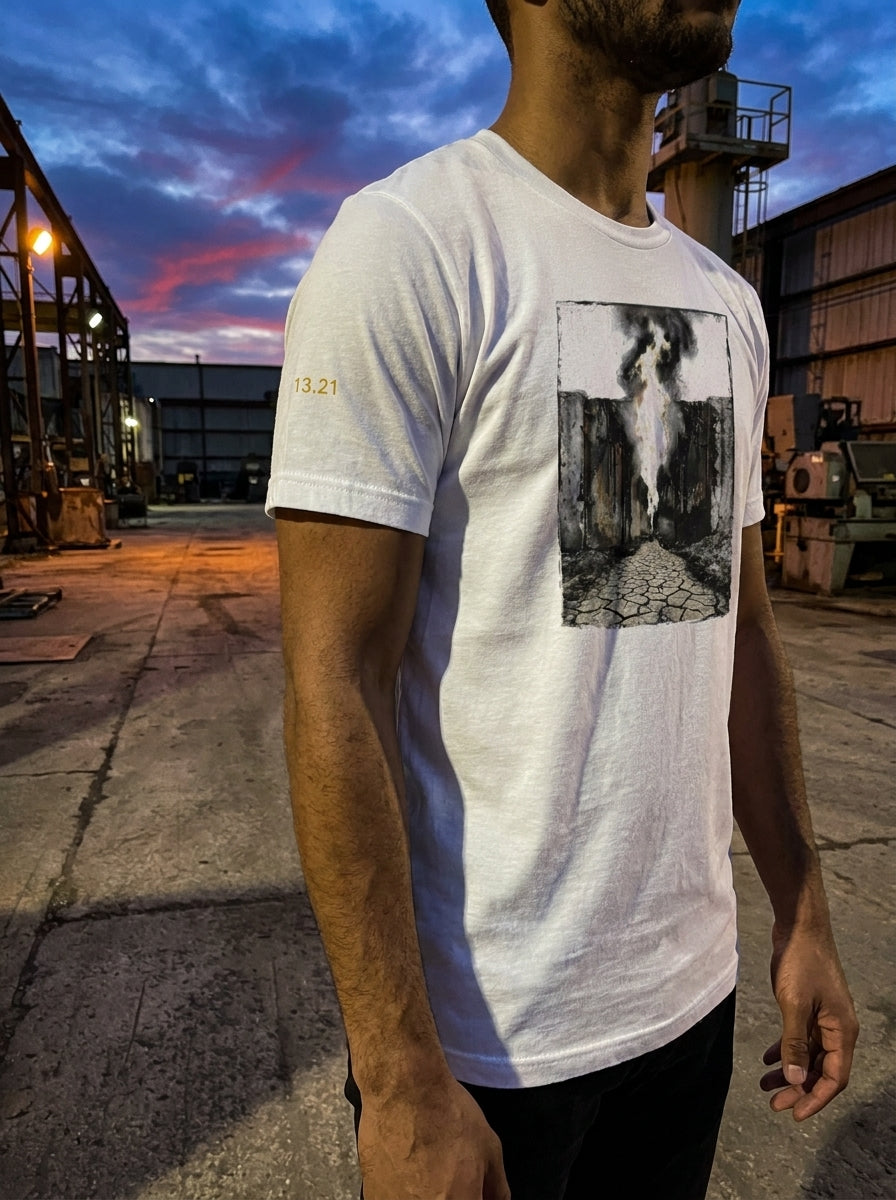 Front-facing vertical lookbook photograph of a model wearing a white premium cotton t-shirt. The garment features a large, centered vertical industrial graphic print depicting a dark column and smog beacon schematic layout. Set against an outdoor landscape background with a dramatic twilight sky showing orange and blue horizon gradients.
