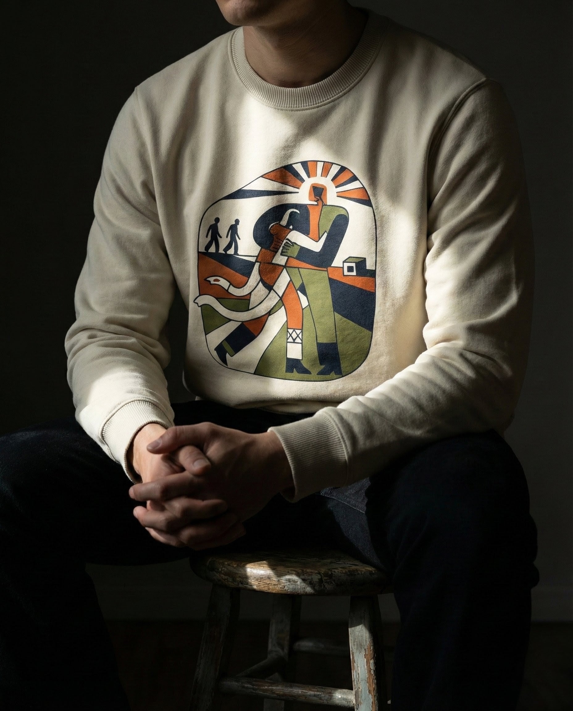 Person wearing a beige sweatshirt with a colorful graphic design sitting on a stool against a dark background