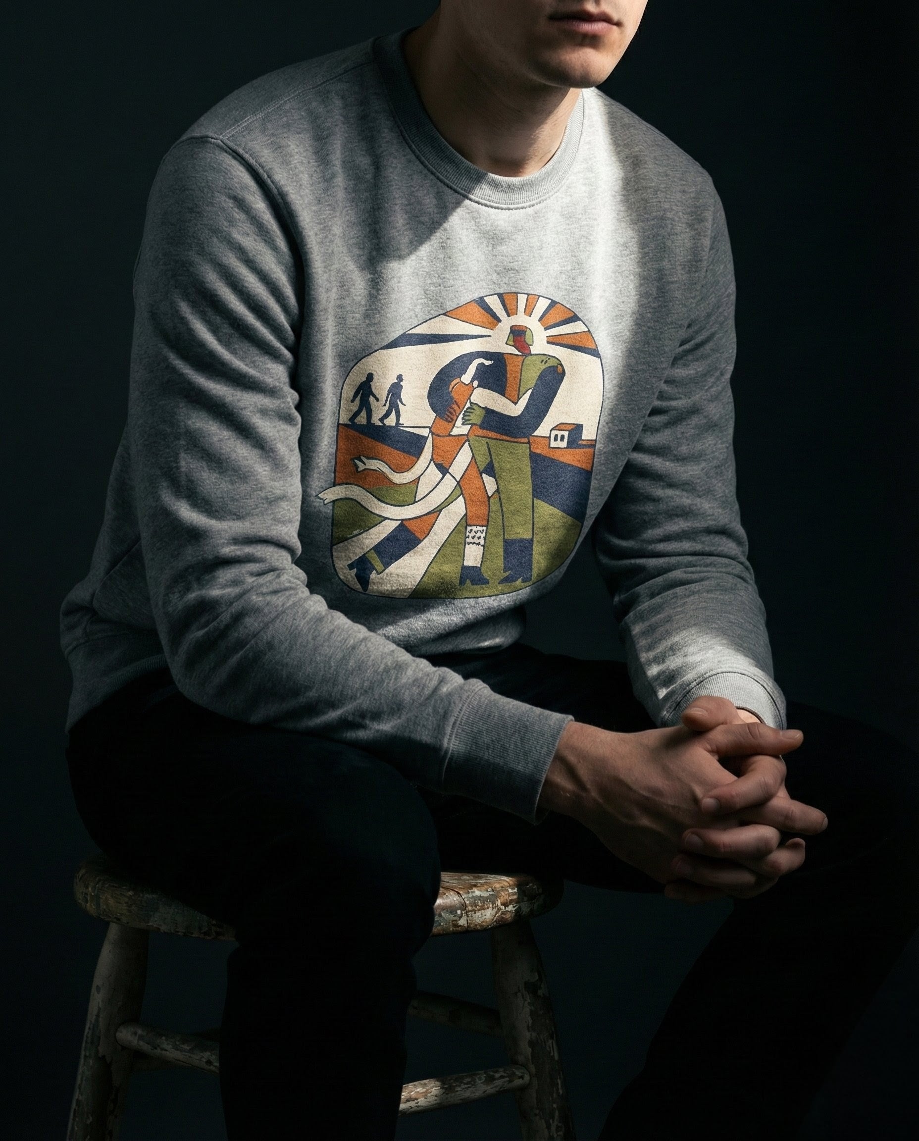 Person wearing a gray sweatshirt with a colorful graphic design sitting on a stool against a dark background