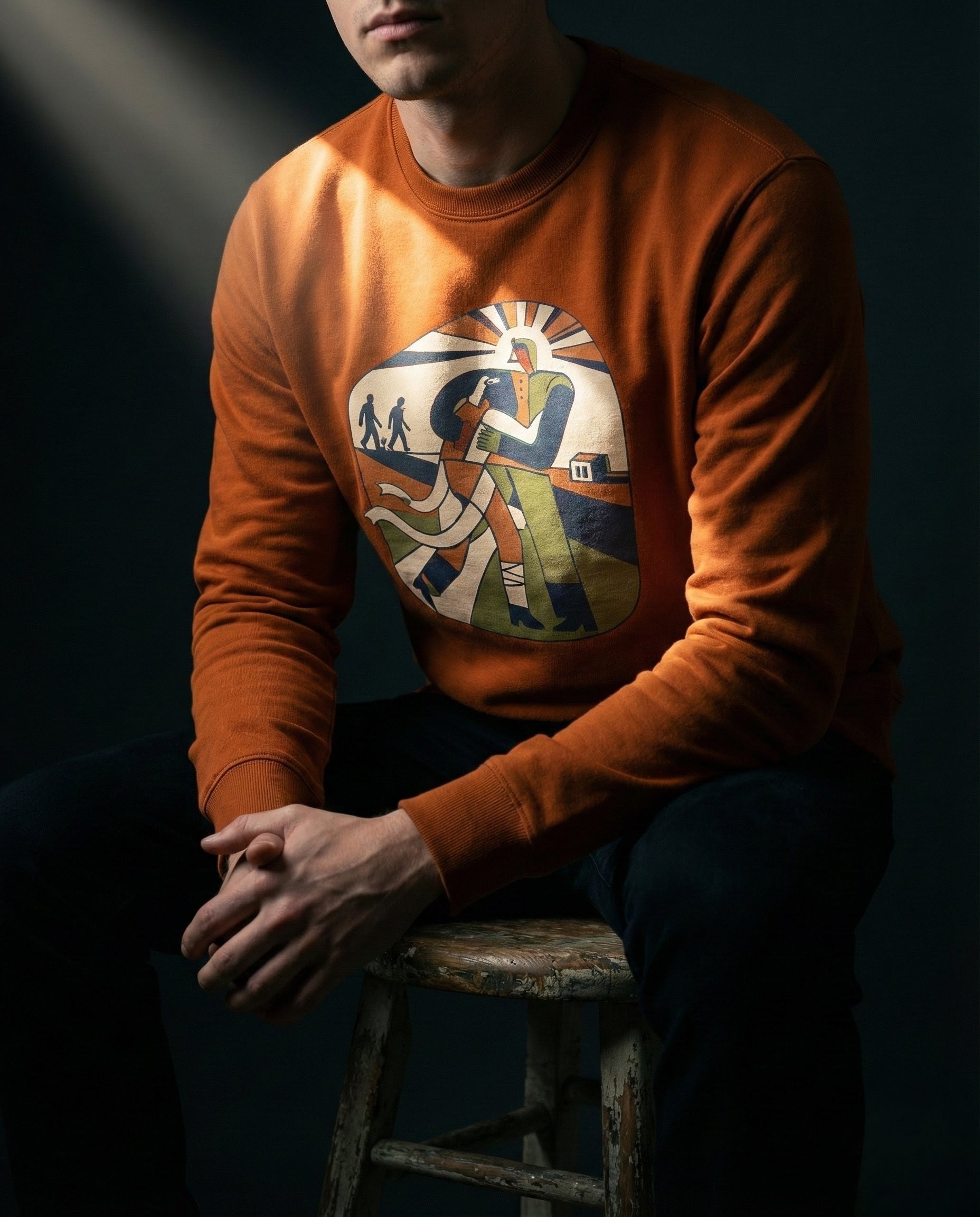 Person wearing an orange sweatshirt with a graphic design, sitting on a stool against a dark background.