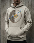 Selah Supply Co. The Rewire Hoodie in Oatmeal Heather. Model wearing a Christian graphic hoodie that visualizes Romans 12:2 through an abstract design of a blue current breaking a rigid grid. Styled against a concrete wall.