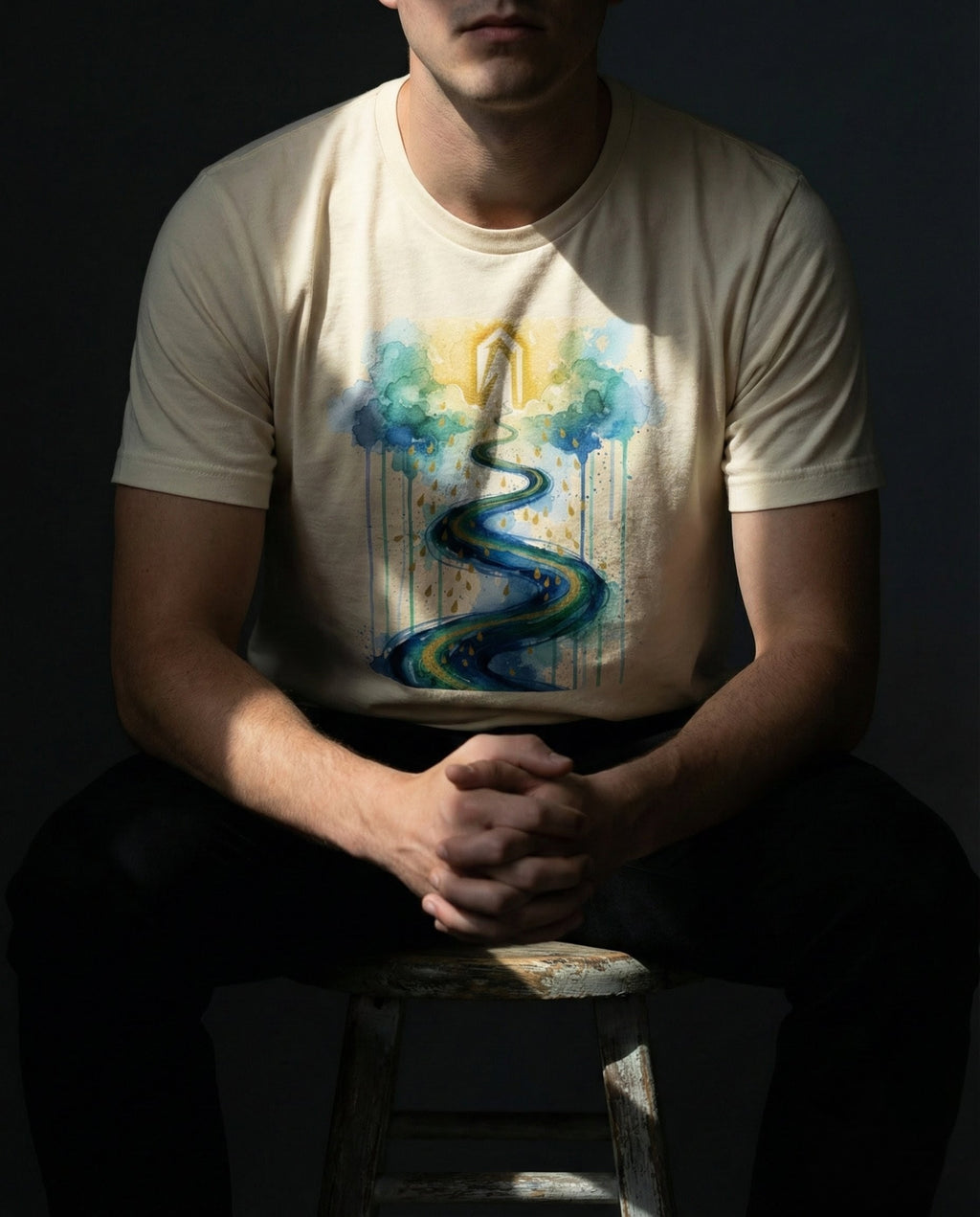 Selah Supply Co. The Impossible Tee in Natural. Model sitting on a wooden stool wearing a Christian graphic tee that visualizes Hebrews 11:6. The design features a golden arch emerging from clouds and a winding river symbolizing the active journey of seeking God.