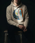Selah Supply Co. The Separation Hoodie in Oatmeal Heather. Model sitting on a wooden stool in a dark workshop wearing a Christian graphic hoodie that visualizes the Parable of the Net from Matthew 13:47 through abstract geometric fish.