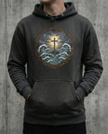 Selah Supply Co. The Trouble Hoodie in Charcoal Heather. Model wearing a Christian graphic hoodie that visualizes John 16:33 through a central cross anchored on rock amidst churning waves and lightning. Styled against a gritty concrete wall.