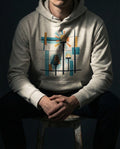 Selah Supply Co. The Bushel Hoodie in Oatmeal Heather. Model wearing a Christian graphic hoodie that visualizes the Parable of the Lamp Stand (Mark 4:21) through a central radiating lamp and a covered lamp within a geometric grid.