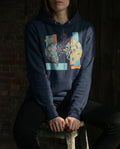 Person wearing a navy blue hoodie with a colorful graphic design, sitting on a wooden stool against a dark background.