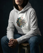 Model wearing a White Cease Striving Hoodie with gold and blue interlocking forms beside scattered black fragments printed on the front.