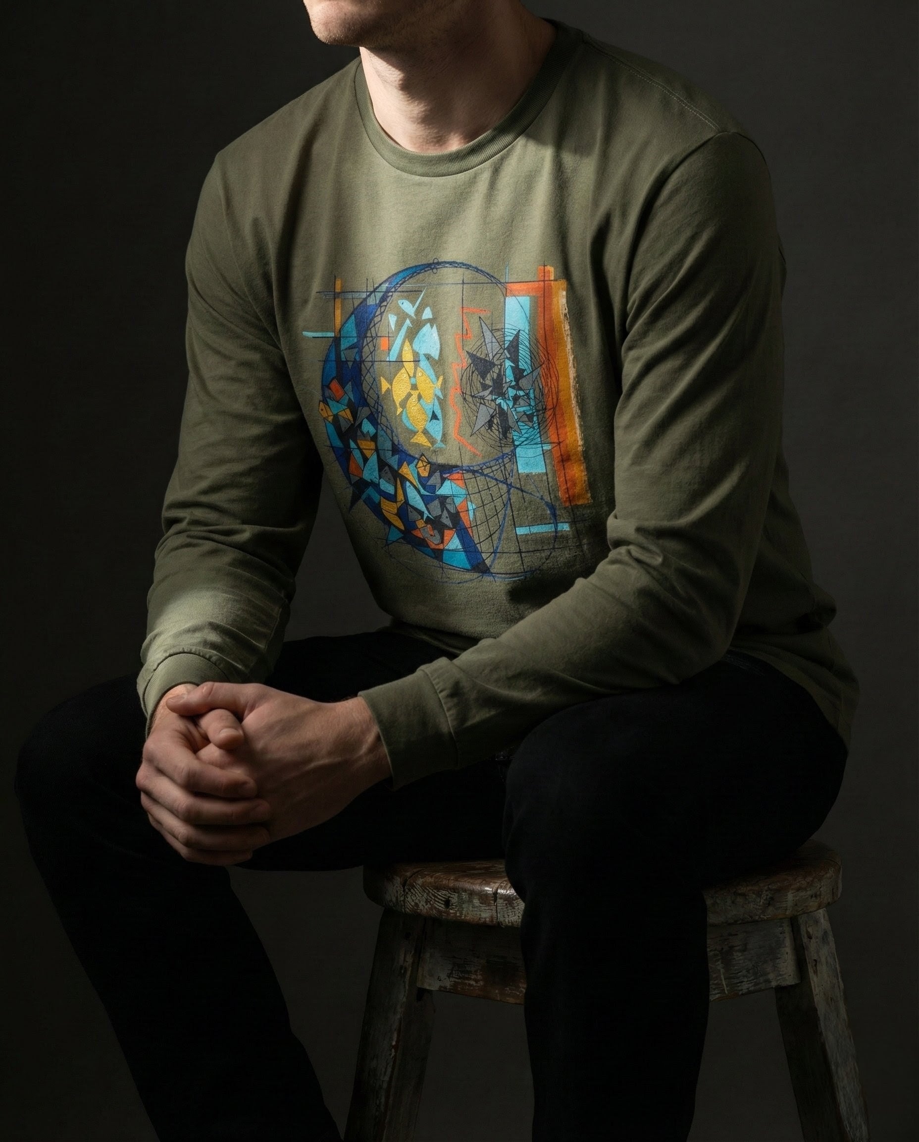 Person wearing a green long-sleeve shirt with a colorful graphic design, sitting on a wooden stool against a dark background.