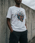 Selah Supply Co. The East West Tee in white. Model standing against a gritty concrete wall wearing a Christian graphic tee that uses a geometric heart and blue wings to visualize the renewal and forgiveness found in Psalm 103.