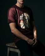 Model seated wearing the Oxblood Black Two Denarii Tee with a central figure supporting a wounded person, flowing white cloth and smaller walking figures.