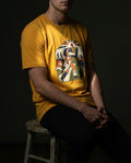 Selah Supply Co. The Two Denarii Tee in Mustard yellow. Model sitting on a wooden stool wearing a Christian graphic tee illustrating the Parable of the Good Samaritan. The design features walking silhouettes and a central embrace, symbolizing active grace.