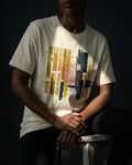"Selah Supply Co. The Tares Tee in Natural. Model sitting on a stool in a technical workshop wearing a graphic tee that uses abstract gold and dark bars to represent the Wheat and Tares from Matthew 13:30."