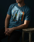 "Selah Supply Co. The Separation Tee in heather deep teal. Model sitting on a wooden stool in a technical workshop wearing a graphic tee that uses abstract geometric fish to represent the sorting of the righteous from Matthew 13:47."