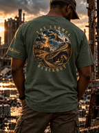A man viewed from behind wearing an olive green t-shirt with a large circular graphic on the back that reads 'THE CARMEL BLACKOUT' and depicts a dramatic desert landscape. He is standing in a ruined city at sunset.