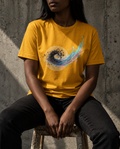 A person seated on a stool wearing a mustard yellow graphic T-shirt with a circular multicolor abstract print on the chest, paired with dark jeans against a textured concrete wall.