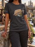 Model wearing a charcoal heather T-shirt with a brass trumpet across a cracked stone tablet, flowing water, and green reeds on the front.