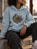 A person wearing a light blue hoodie with a large multicolor geometric and abstract graphic on the front, seated outdoors on stone steps with a city skyline in the background.