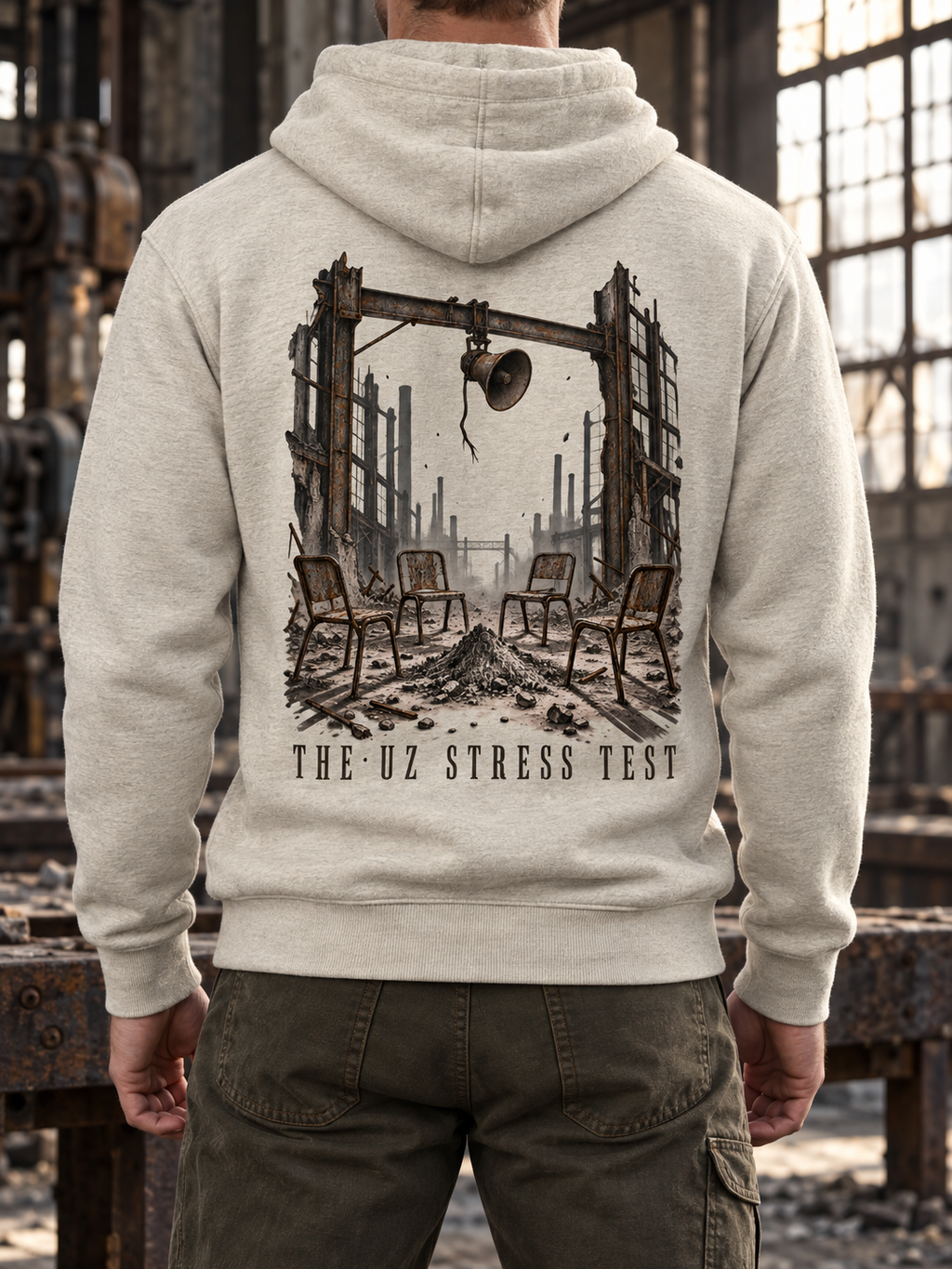 A person shown from the back wearing a light heather gray hoodie with a large dark graphic print of a ruined industrial scene and the text 'THE UZ STRESS TEST' on the back, standing in a dilapidated factory interior.