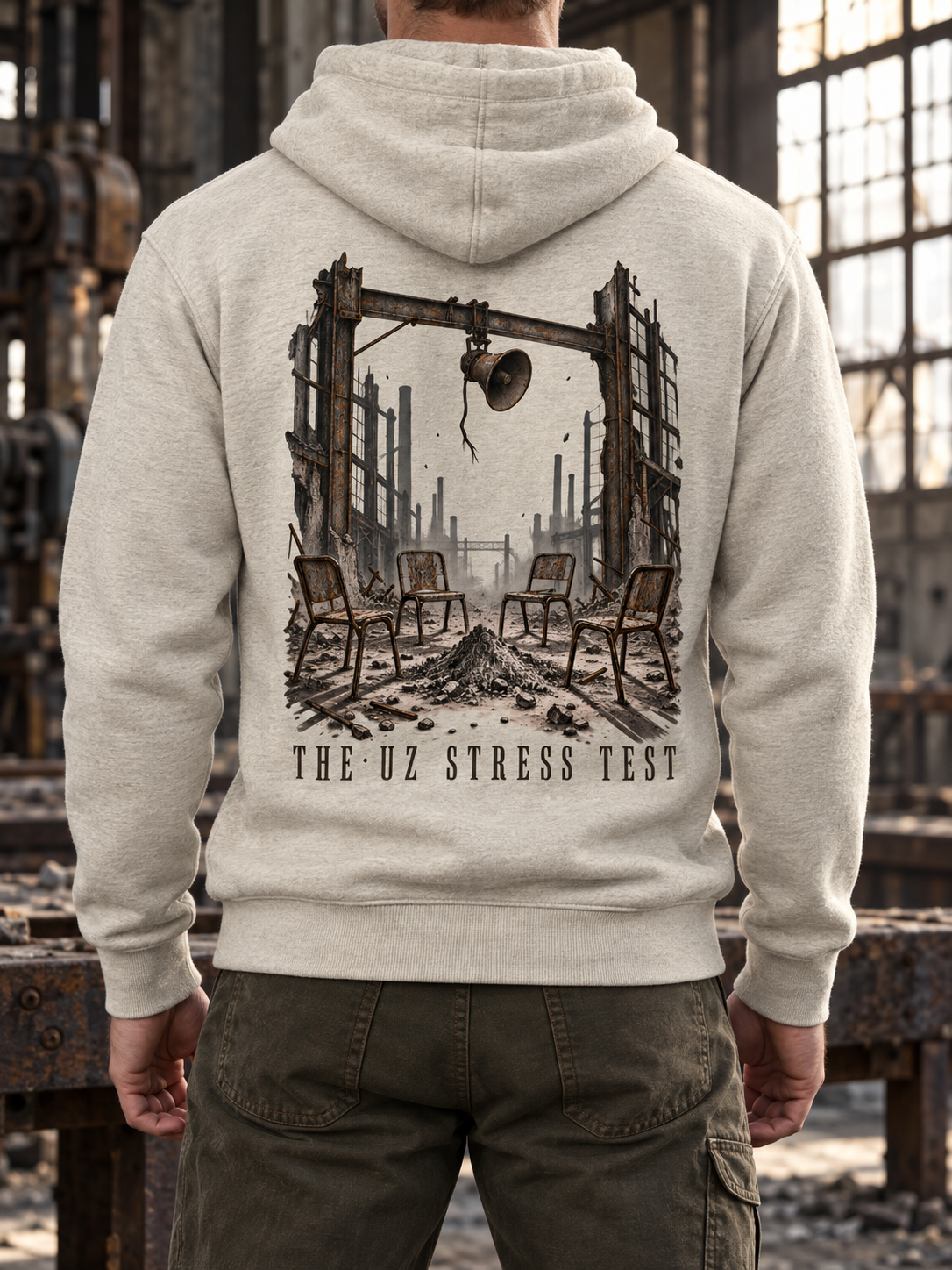 A person shown from the back wearing a light heather gray hoodie with a large dark graphic print of a ruined industrial scene and the text 'THE UZ STRESS TEST' on the back, standing in a dilapidated factory interior.