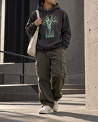 Model wearing the Charcoal Heather Thirty Sixty Hundred Hoodie with a tall teal, navy and gold spray of seeds and leaves above small seedlings.
