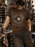 Front view of a standing model in a workshop, wearing a heather brown T-shirt with a white reaction-chamber schematic surrounding a bright central spark centered on the front.