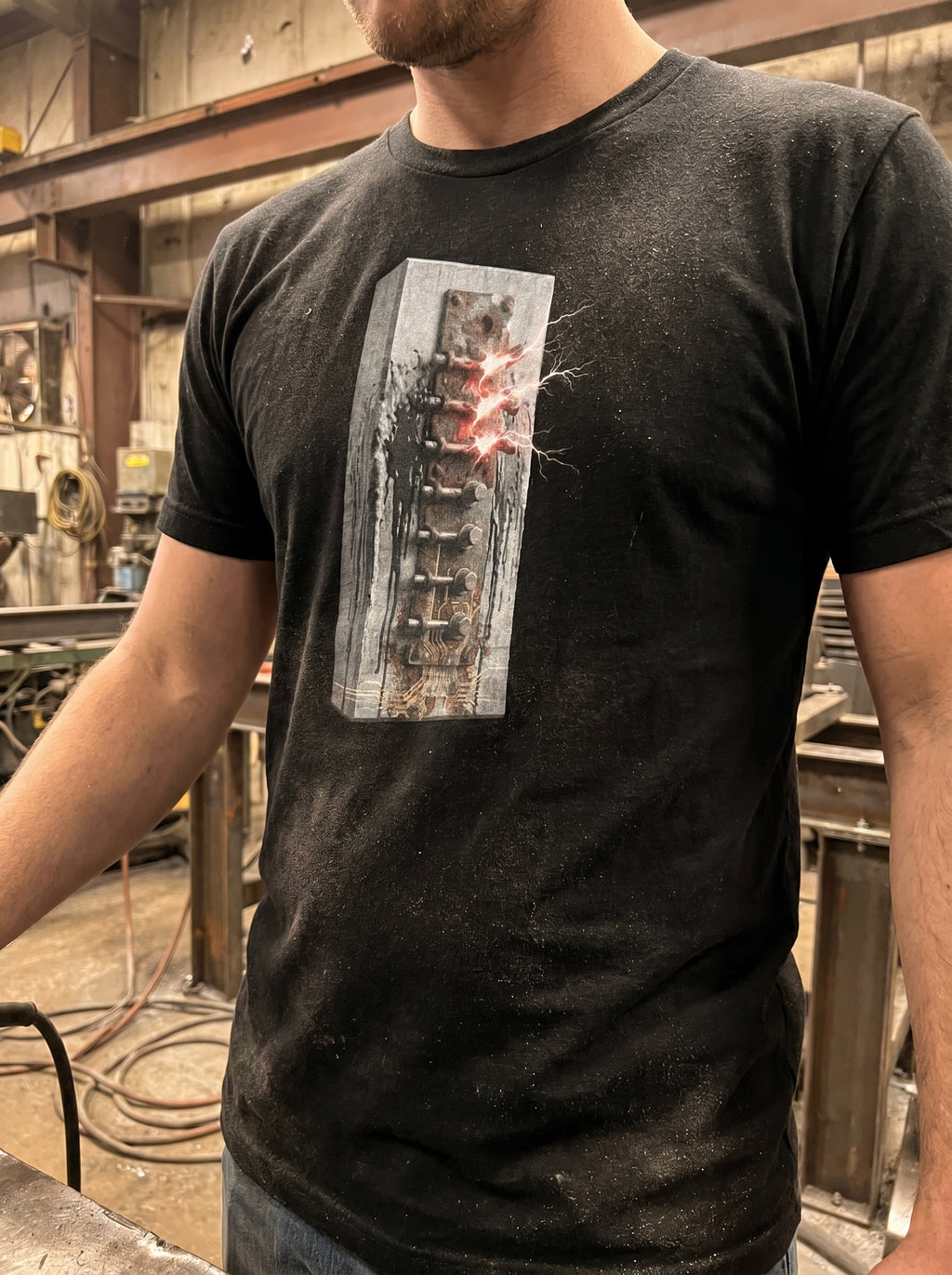 Rear-angle outdoor lookbook photograph of a model wearing the dark grey heather industrial t-shirt. The shot highlights the vertical orientation of the technical mechanical spine graphic running down the back, captured against a raw, weathered concrete architectural wall under natural daylight.