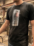 Rear-angle outdoor lookbook photograph of a model wearing the dark grey heather industrial t-shirt. The shot highlights the vertical orientation of the technical mechanical spine graphic running down the back, captured against a raw, weathered concrete architectural wall under natural daylight.